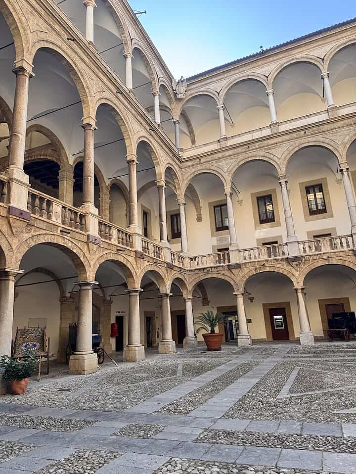 Stone courtyard with arches, columns, potted plants, and balconies inside a historic building.