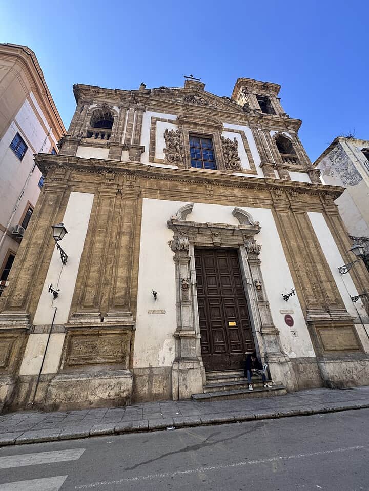A person sits on steps outside an ornate church with tall columns and intricate carvings under a clear blue sky.