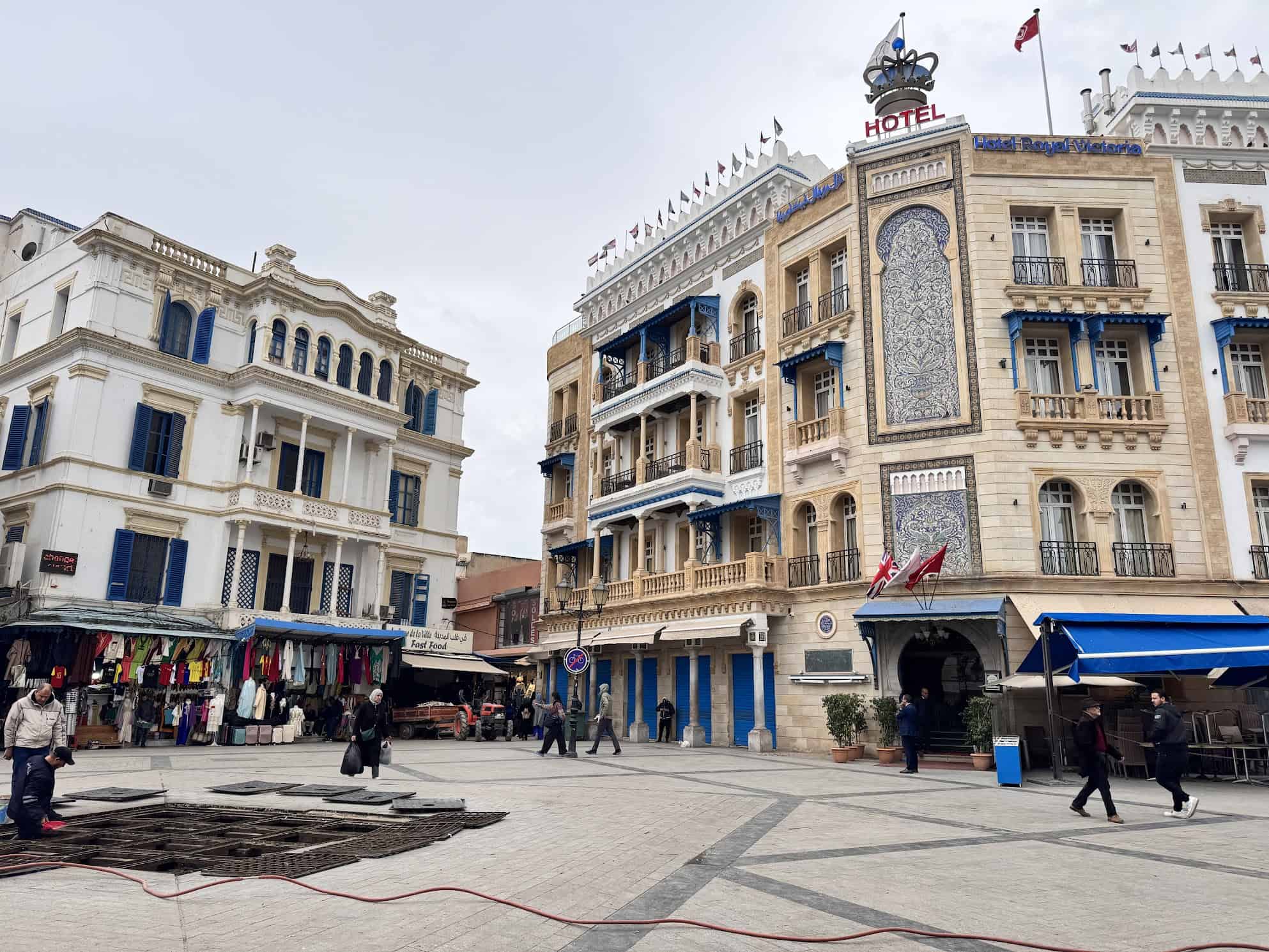 People walk in a plaza with historic buildings, shops, and a hotel decorated with blue and white details.