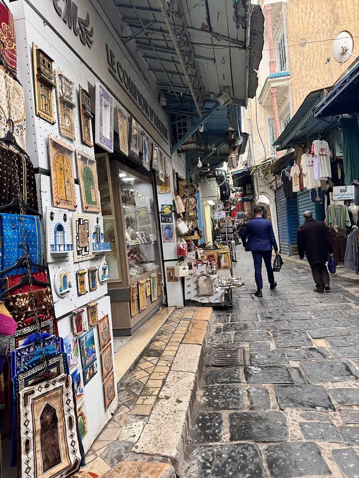 Narrow cobblestone street with shops selling souvenirs, rugs, and bags; two men walk away.