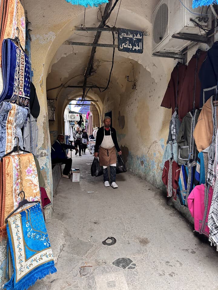 Narrow alleyway market with colorful fabrics, people walking, and archways overhead in a Middle Eastern town.