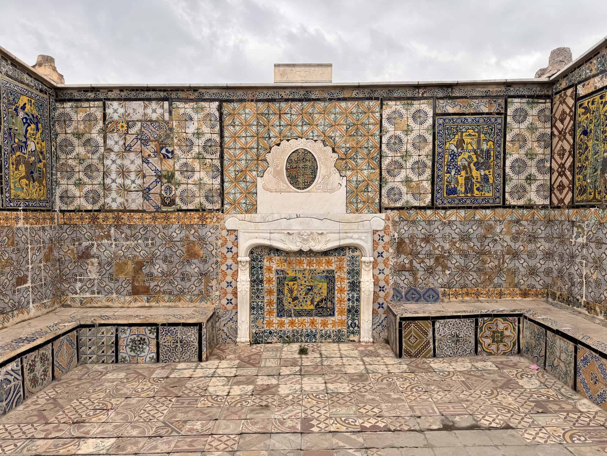 Mosaic-tiled courtyard with a decorative marble fireplace, colorful patterns cover walls, floor, and benches.