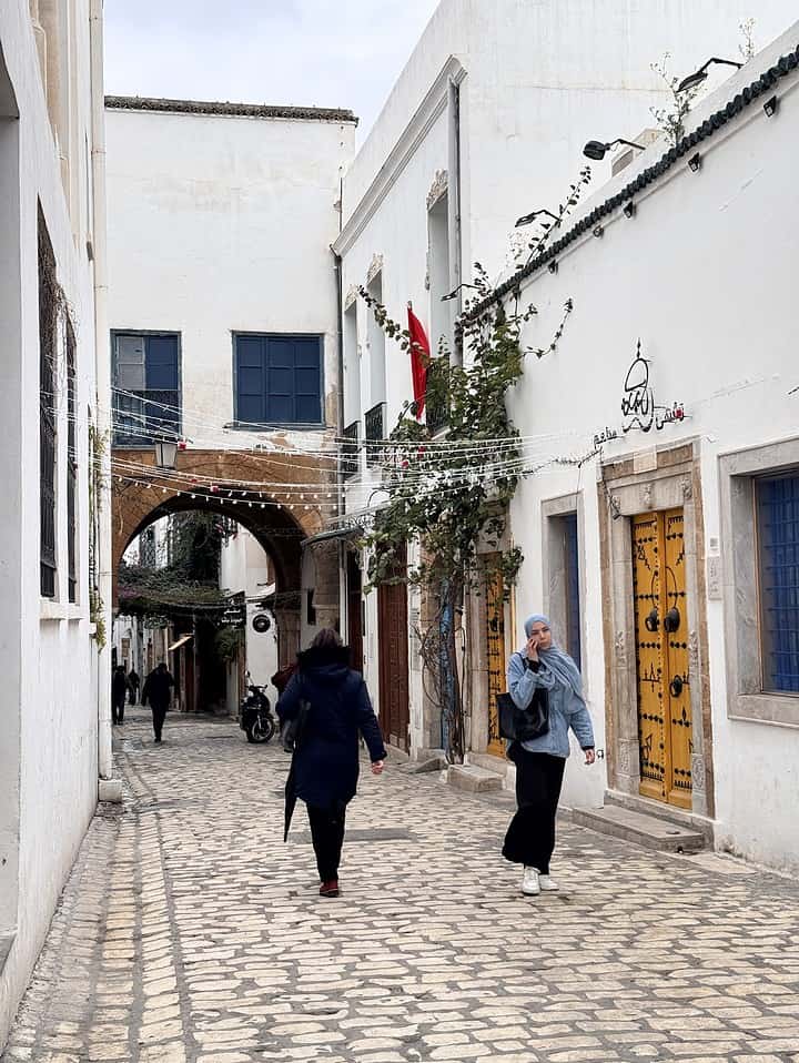People walk down a cobblestone street between white buildings with colorful doors and hanging lights.