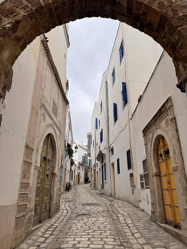 A cobblestone street with white buildings and colorful doors, viewed through a stone archway.