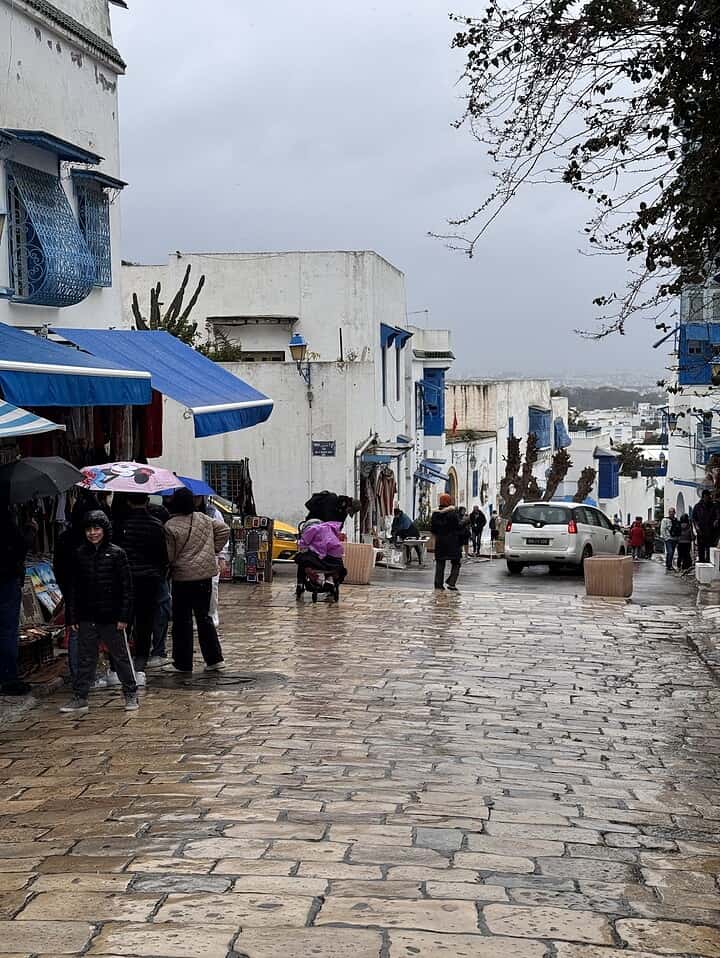 People walk on a rainy, cobblestone street lined with white buildings and blue accents; some hold umbrellas.