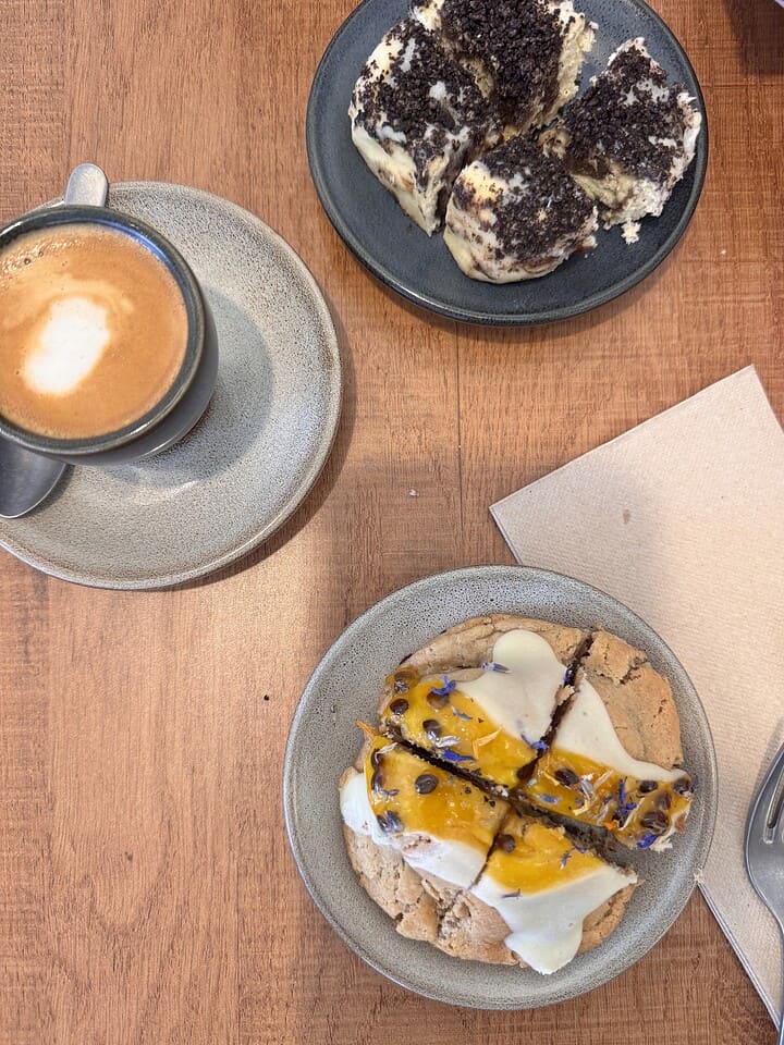 A cup of coffee, a frosted cookie, and a plate of chocolate cake slices on a wooden table.