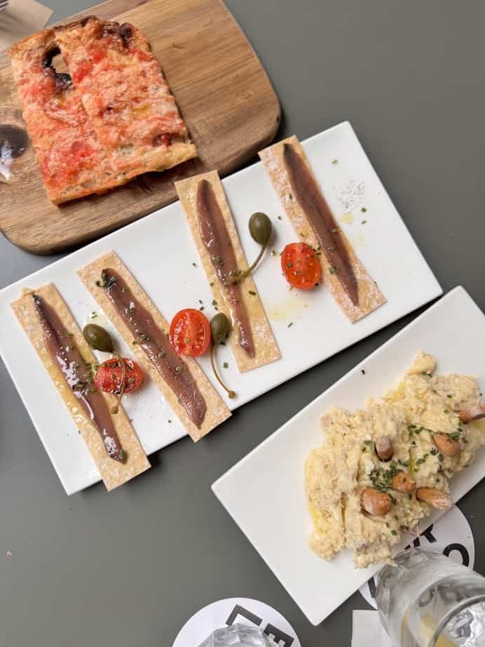Rectangular crackers with anchovies, a pizza slice, and dip with nuts and herbs on plates—perfect for those seeking fresh food inspired by Barcelona markets.