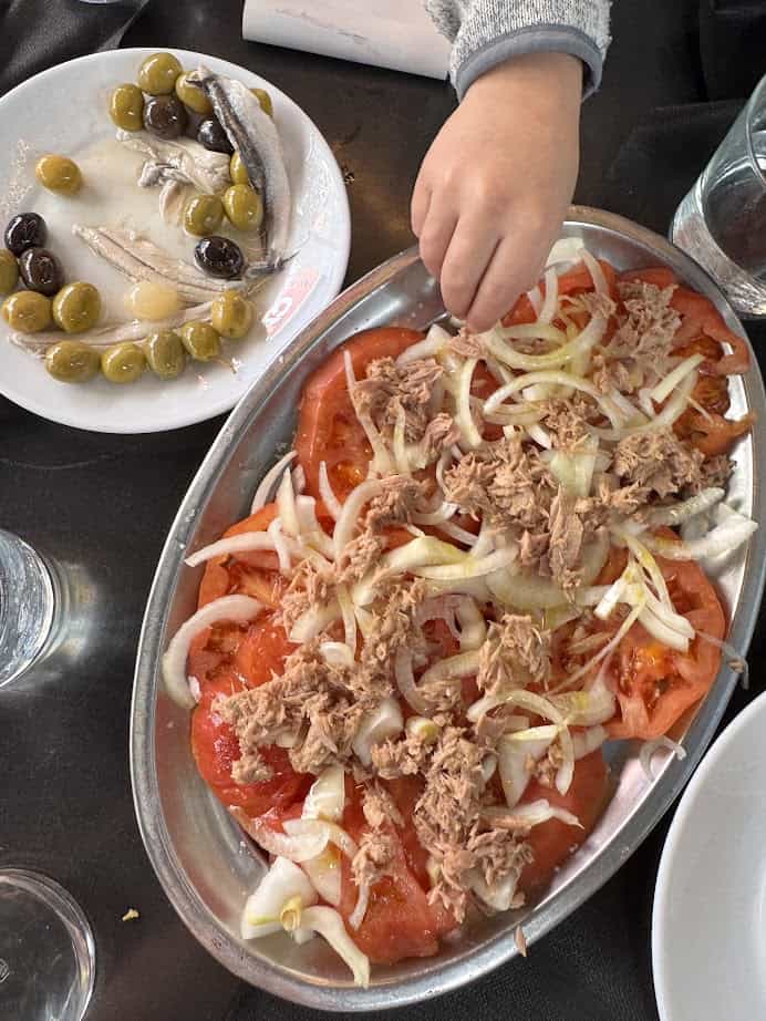 A child&rsquo;s hand reaches for a tomato, onion, and tuna salad beside a plate of olives and anchovies&mdash;fresh food inspired by the flavors found in Barcelona markets.