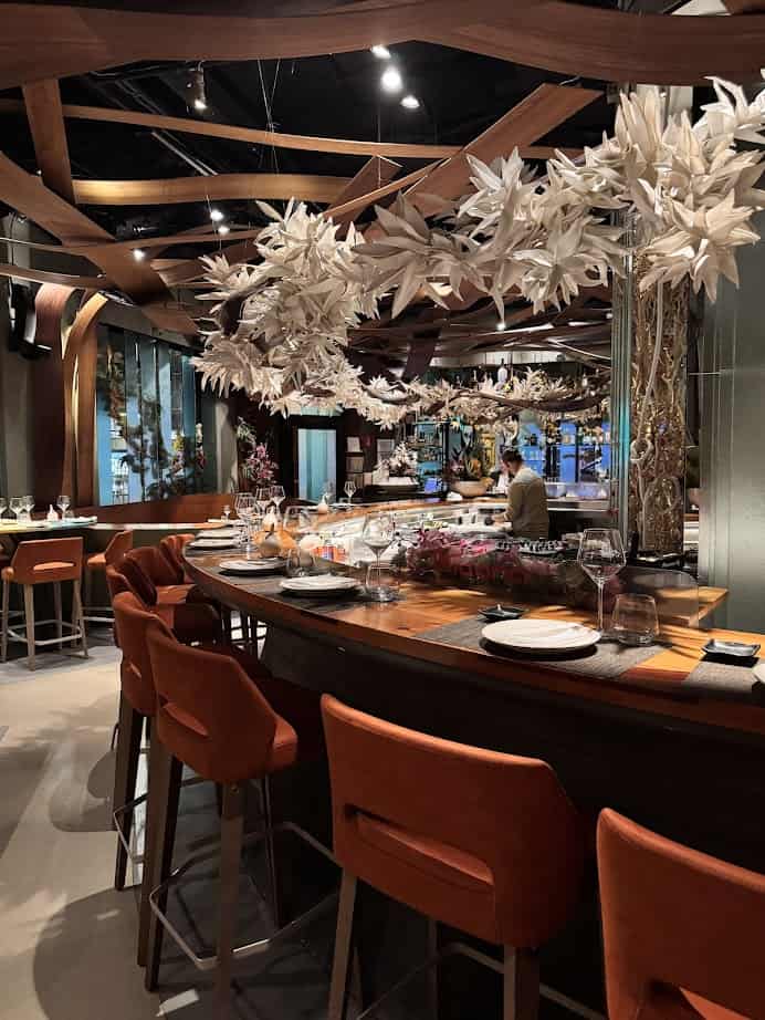 A modern restaurant with orange bar stools, wooden accents, and elaborate white floral ceiling decorations—an ideal spot where to eat fresh food inspired by Barcelona markets.
