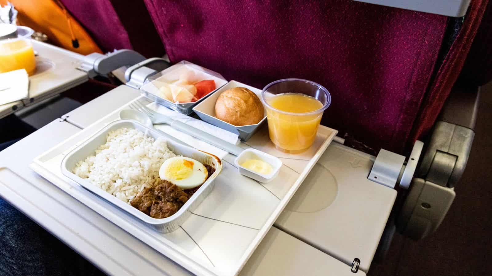 Airplane meal with rice, egg, meat, bread, fruit, butter, and orange juice on a tray table.