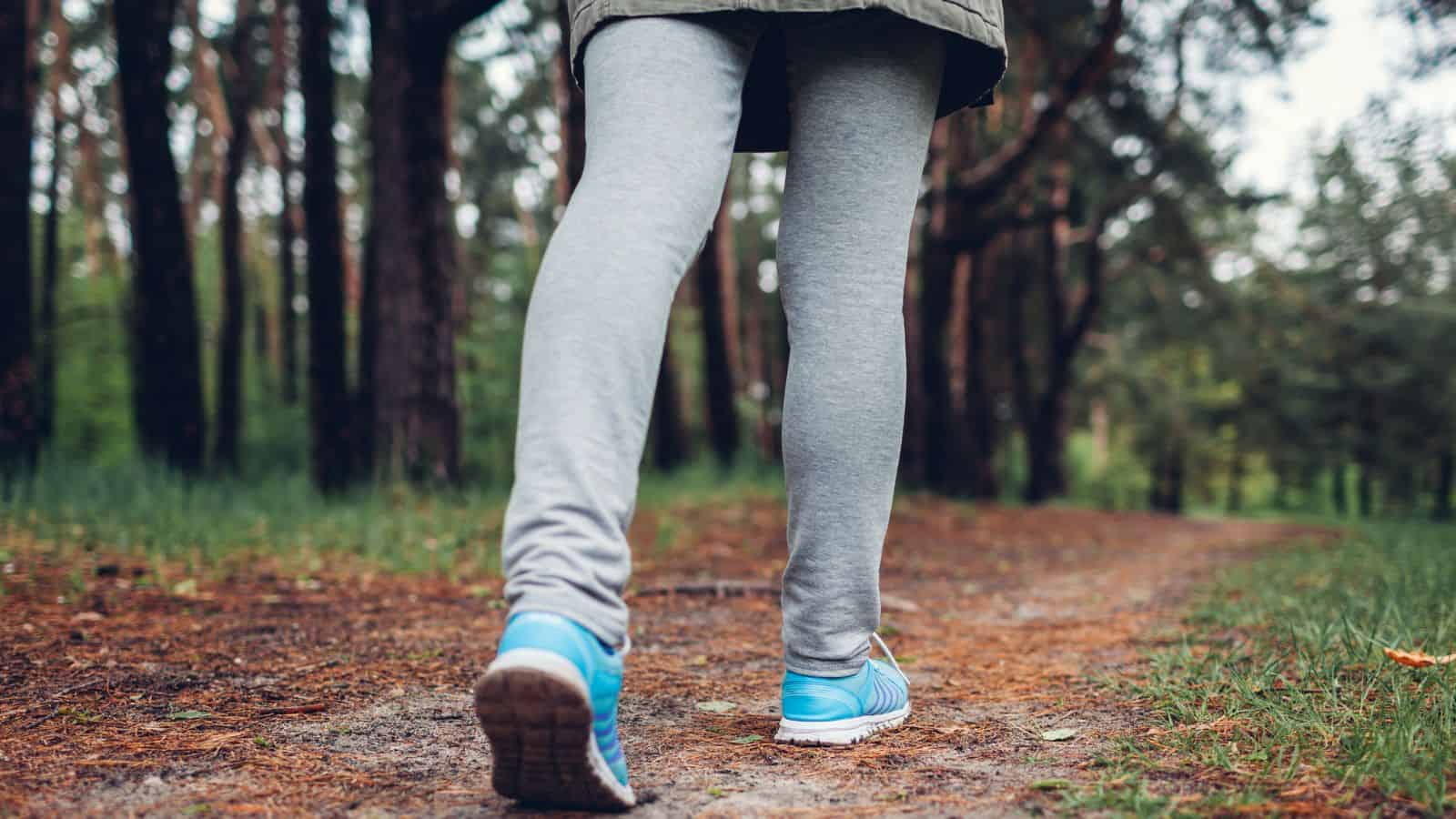 Person wearing gray leggings and blue sneakers walking on a forest trail, surrounded by trees and greenery.