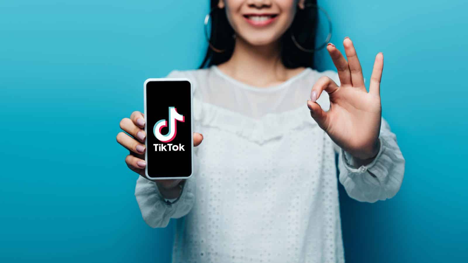 Smiling woman holds a phone with the TikTok logo and makes an OK hand gesture against a blue background.