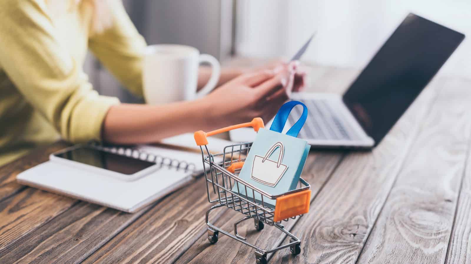 A person works on a laptop with a notebook, while a gift bag sits in a small shopping cart in the foreground.