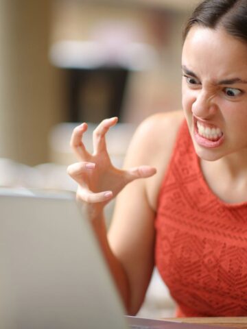 A frustrated woman in an orange top glares at her laptop, clenching her hands in anger beside a coffee cup.