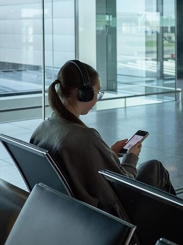 A woman with headphones sits in an empty airport terminal, looking at her phone with luggage beside her.