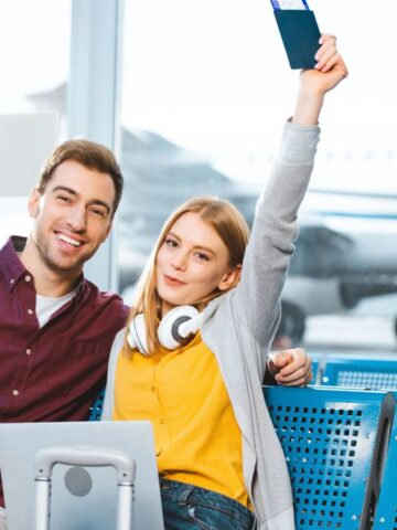 Smiling couple holding passports and tickets, sitting in an airport waiting area with a plane outside window.