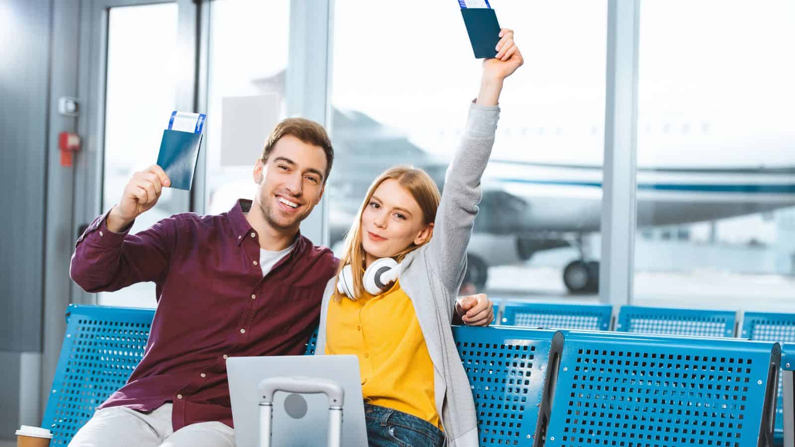 Smiling couple holding passports and tickets, sitting in an airport waiting area with a plane outside window.
