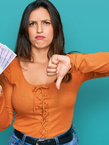 Woman holding cash with one hand and showing a thumbs down with the other, looking dissatisfied.