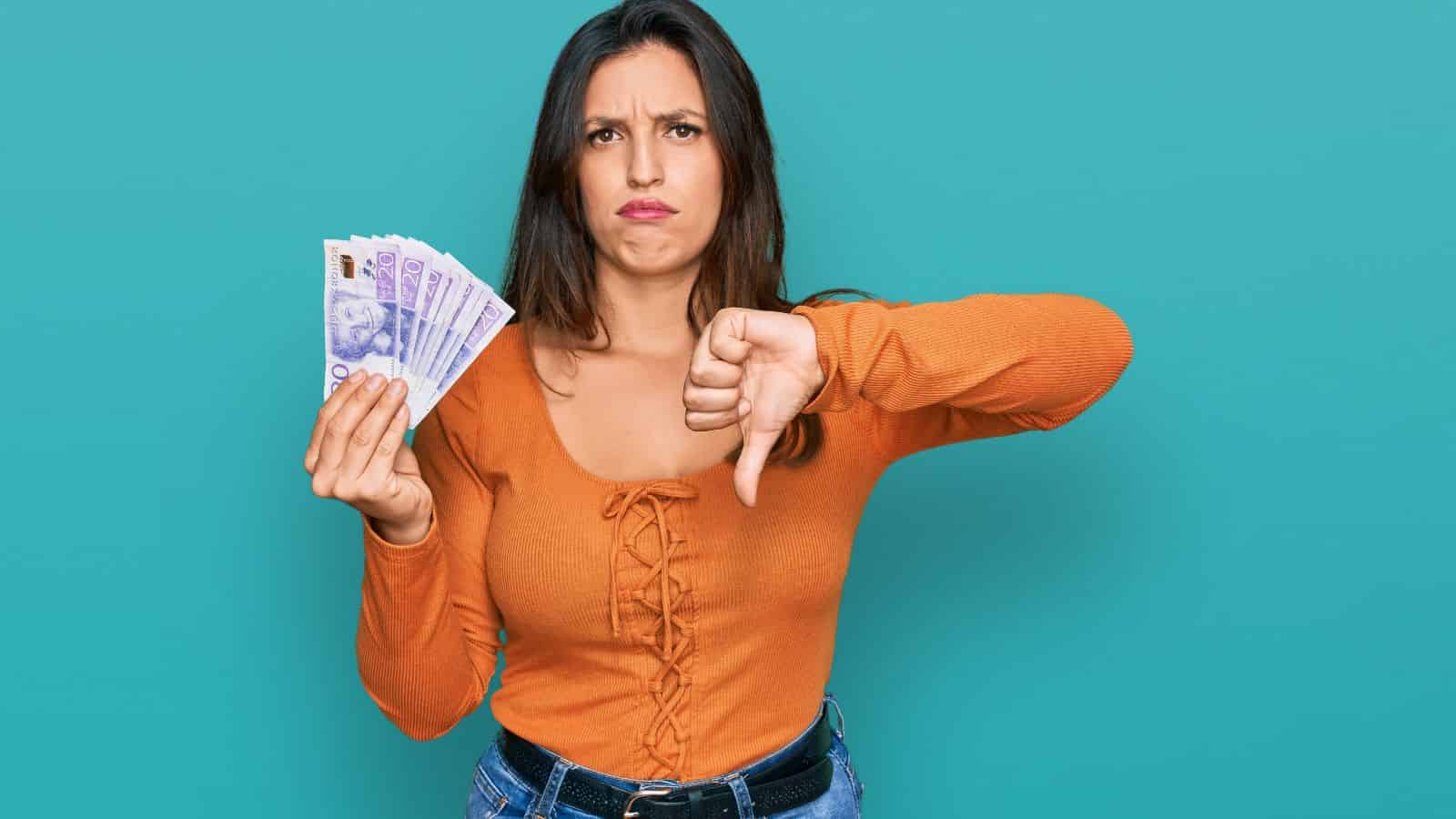 Woman holding cash with one hand and showing a thumbs down with the other, looking dissatisfied.