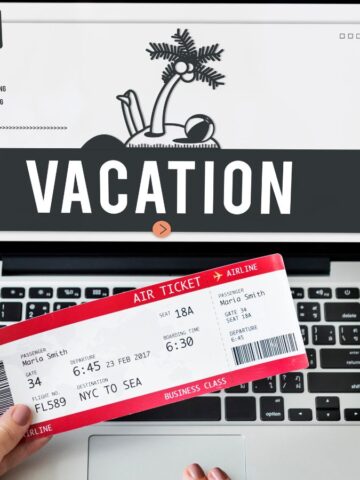 A hand holds a plane ticket in front of a laptop displaying "VACATION" on the screen, ready to book with airline points and discover new travel tips.