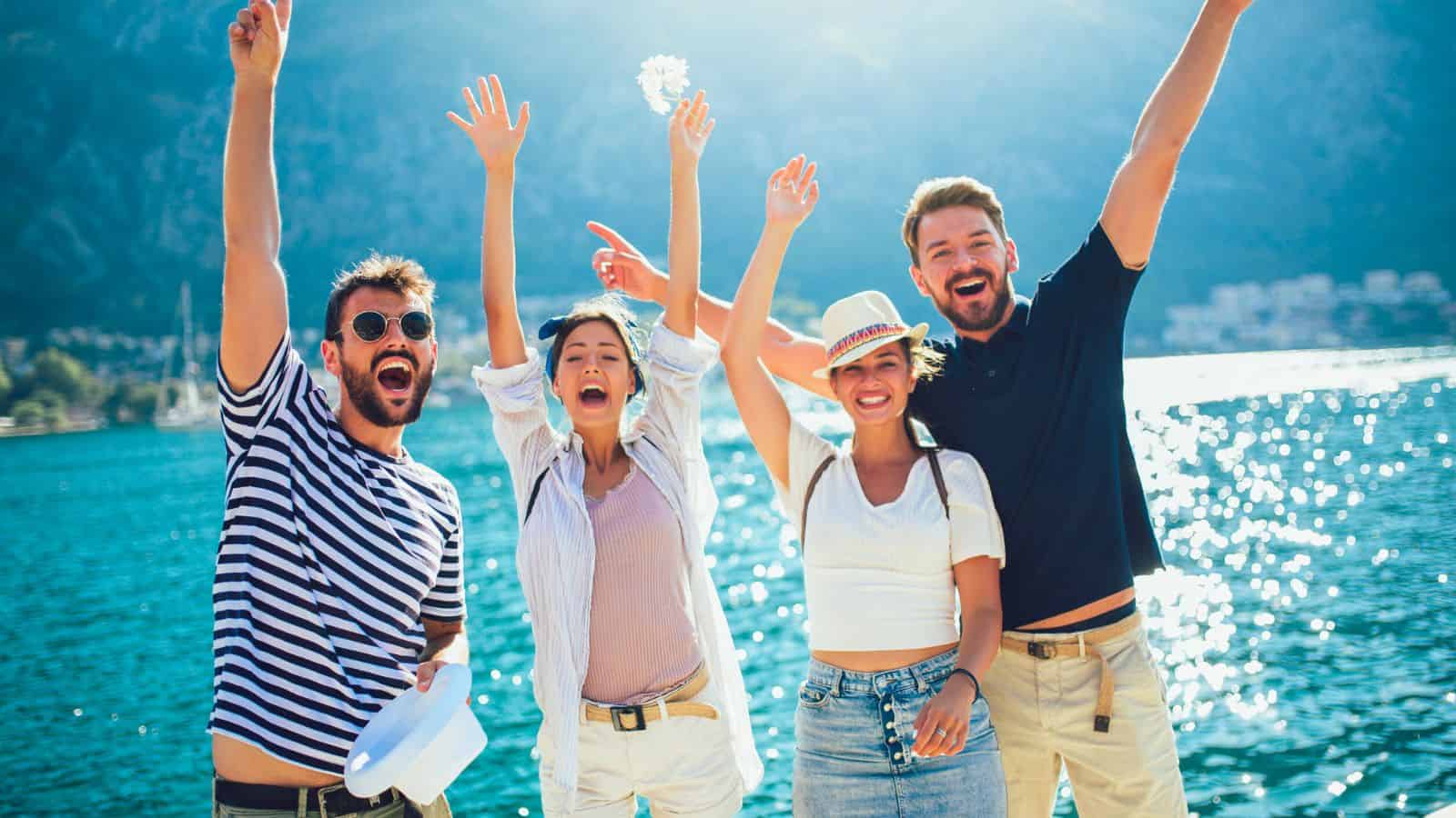 Four friends smiling and raising their arms, standing by a bright blue lake with mountains in the background.