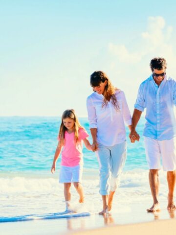 A family of four walks along the beach shore on a sunny day, holding hands and smiling.
