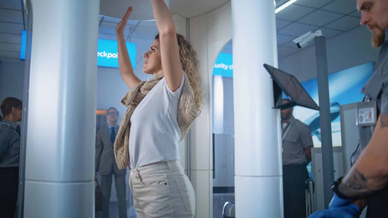 Woman stands with arms raised inside airport security scanner; security personnel and travelers are nearby.