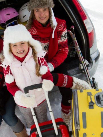 Three people in winter clothes sit in a car trunk with sleds, suitcases, and ski gear on snow.