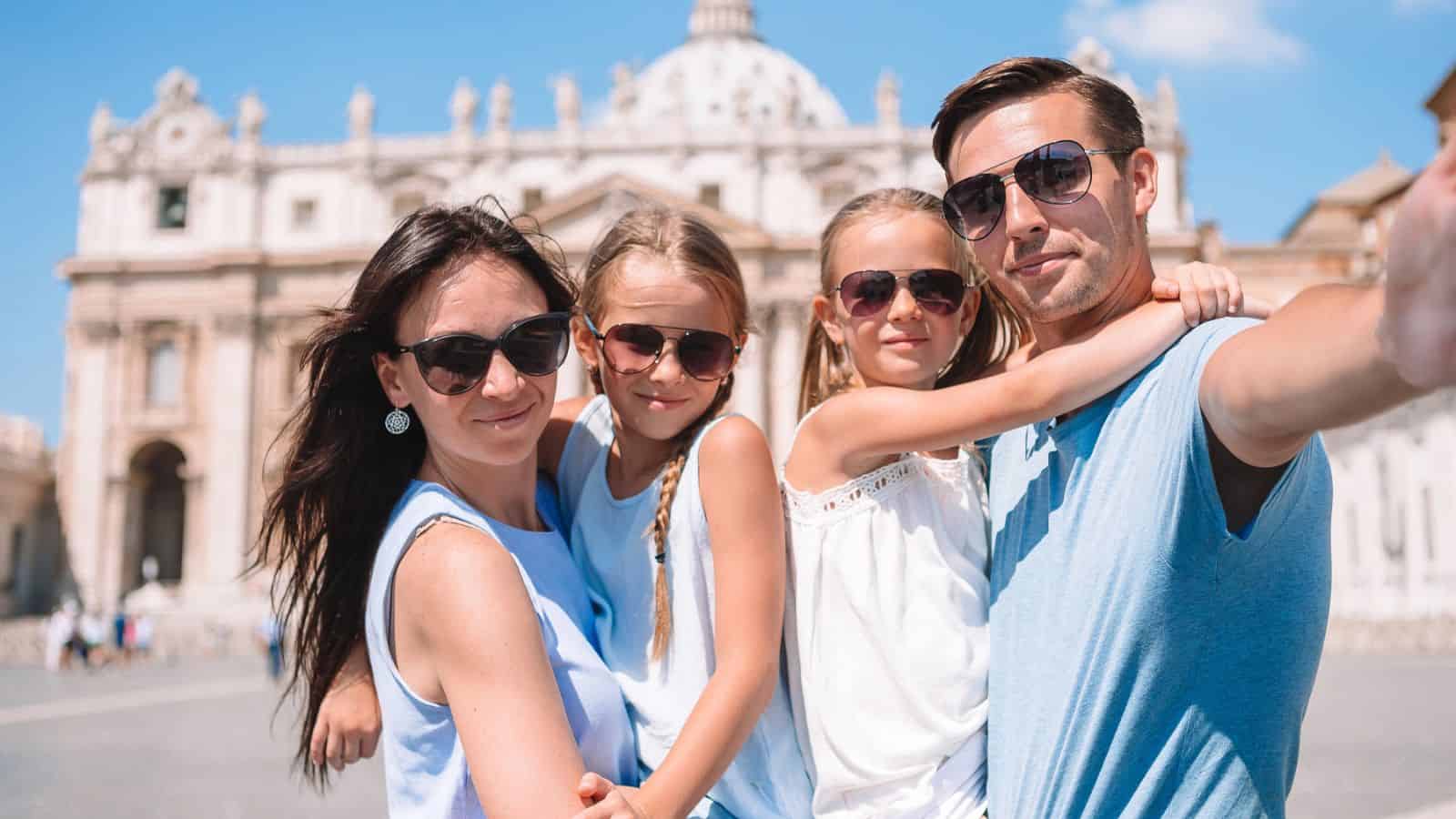 A family of four wearing sunglasses takes a selfie in front of a historic building on a sunny day.