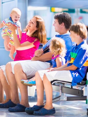 A family with three children sits in an airport, smiling with luggage and colorful toys beside them.