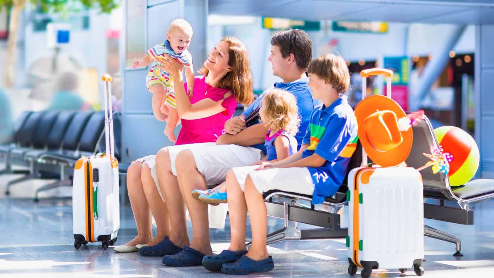 A family with three children sits in an airport, smiling with luggage and colorful toys beside them.