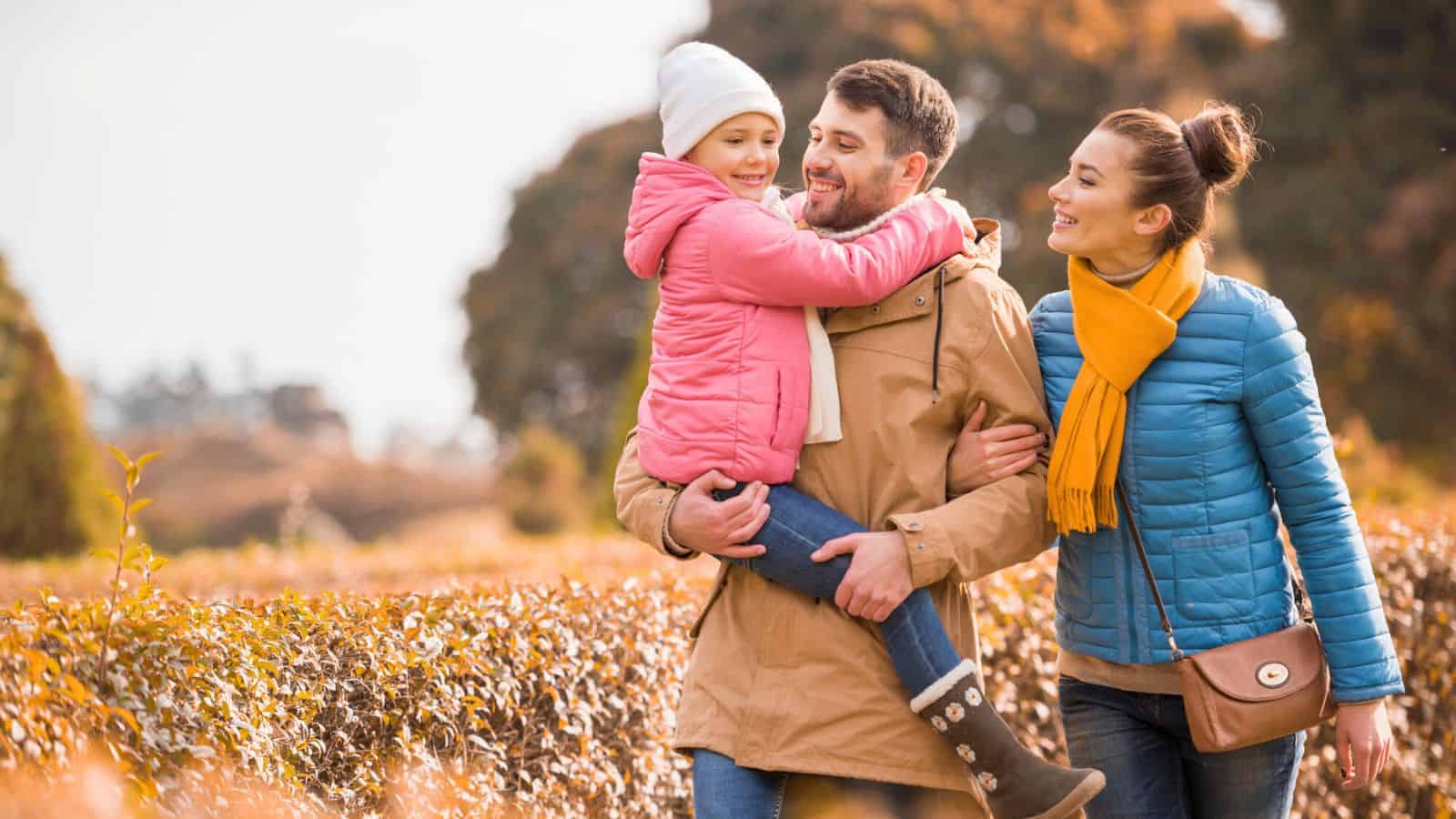 A family of three walks outdoors in autumn, smiling and enjoying the fall scenery.