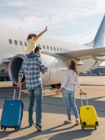 A family with suitcases walks toward an airplane on the tarmac; a child waves while on an adult's shoulders.