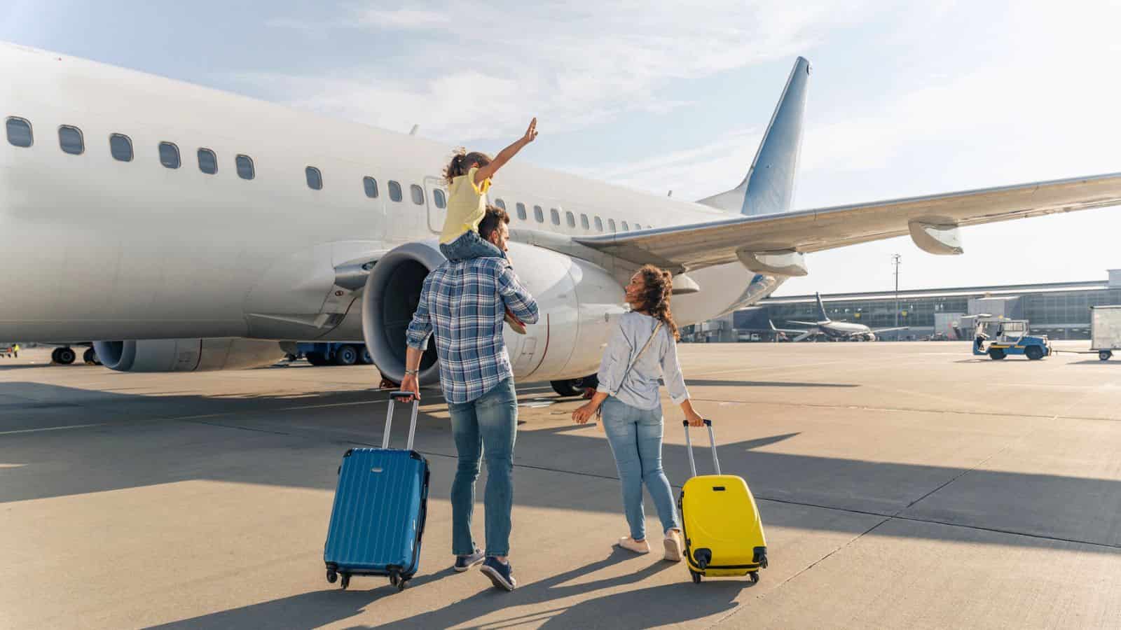 A family with suitcases walks toward an airplane on the tarmac; a child waves while on an adult's shoulders.