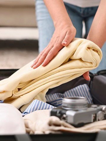 Person packing a suitcase, placing a yellow sweater on top of neatly folded clothes and travel essentials.