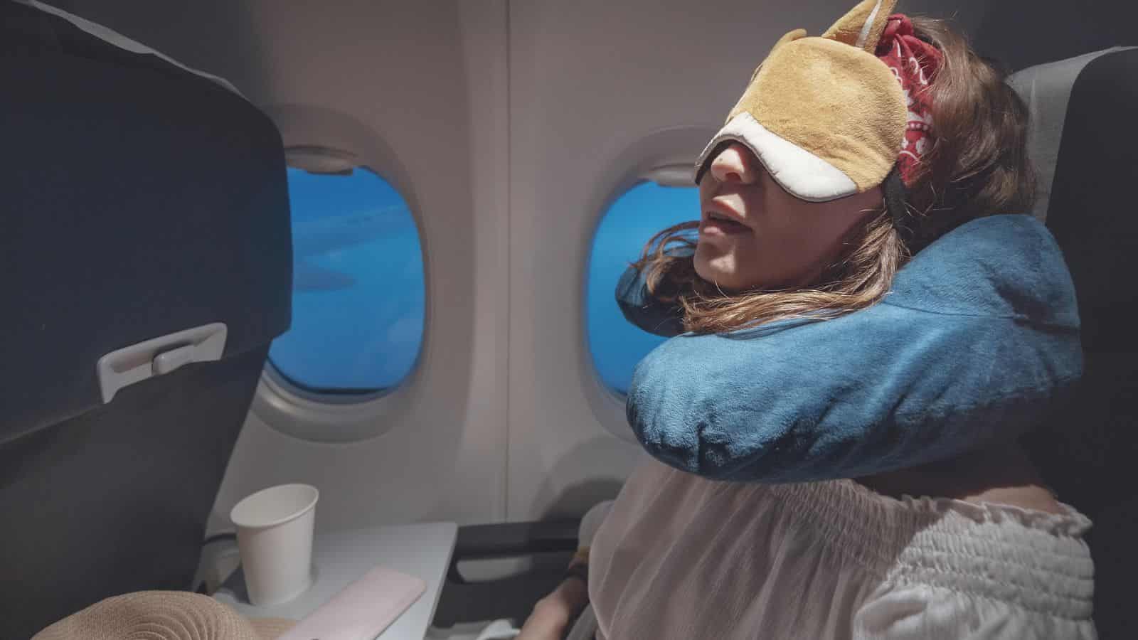 A woman demonstrates the comfort of travel products, sleeping on a plane with a neck pillow and eye mask beside the window, her tray holding a cup.