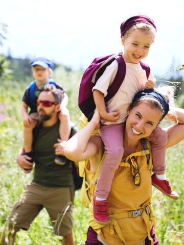 A family hikes through a sunny field, parents carrying young children on their shoulders, all smiling.