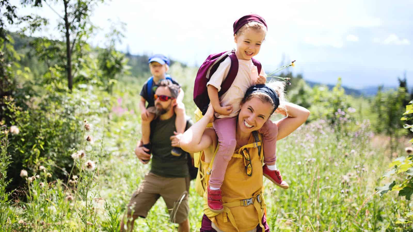 A family hikes through a sunny field, parents carrying young children on their shoulders, all smiling.