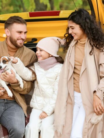 A smiling family with a dog sits on a car trunk in a park, surrounded by autumn leaves and trees.