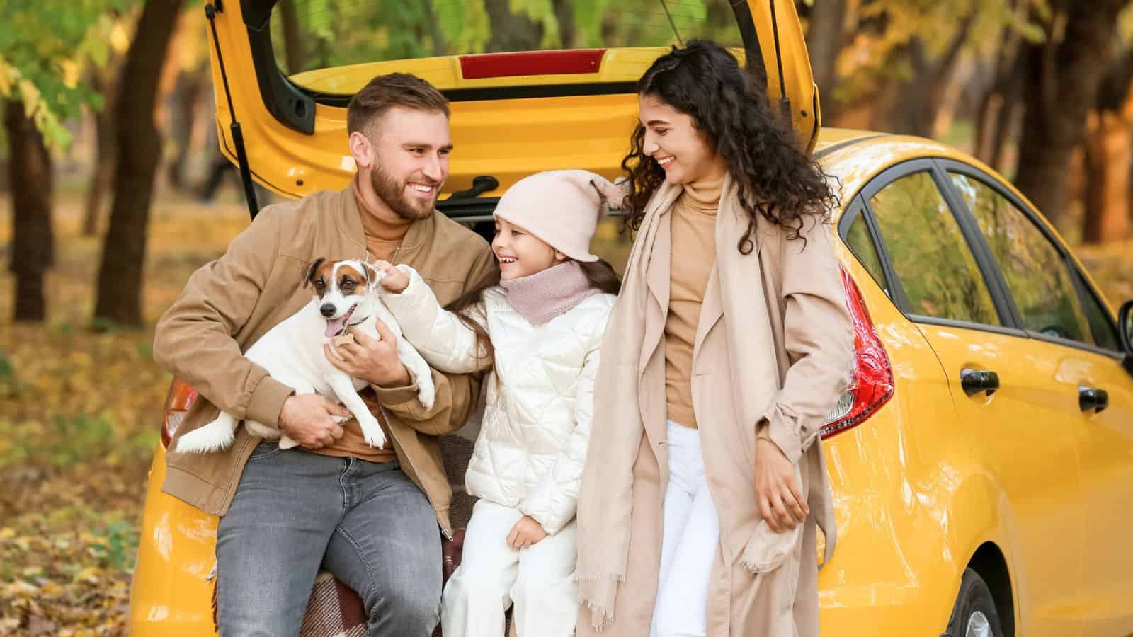A smiling family with a dog sits on a car trunk in a park, surrounded by autumn leaves and trees.