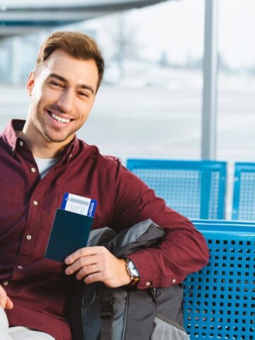 Smiling man at airport holding a passport and boarding pass, sitting on blue seats with a backpack.