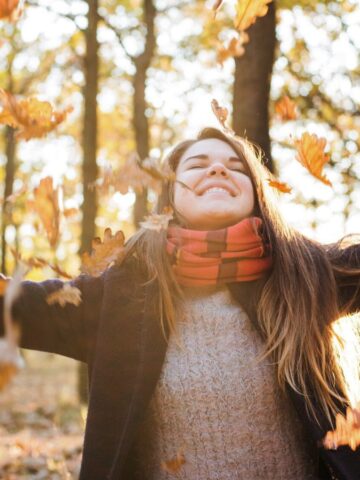 Woman smiling and throwing autumn leaves in the air in a sunlit forest.