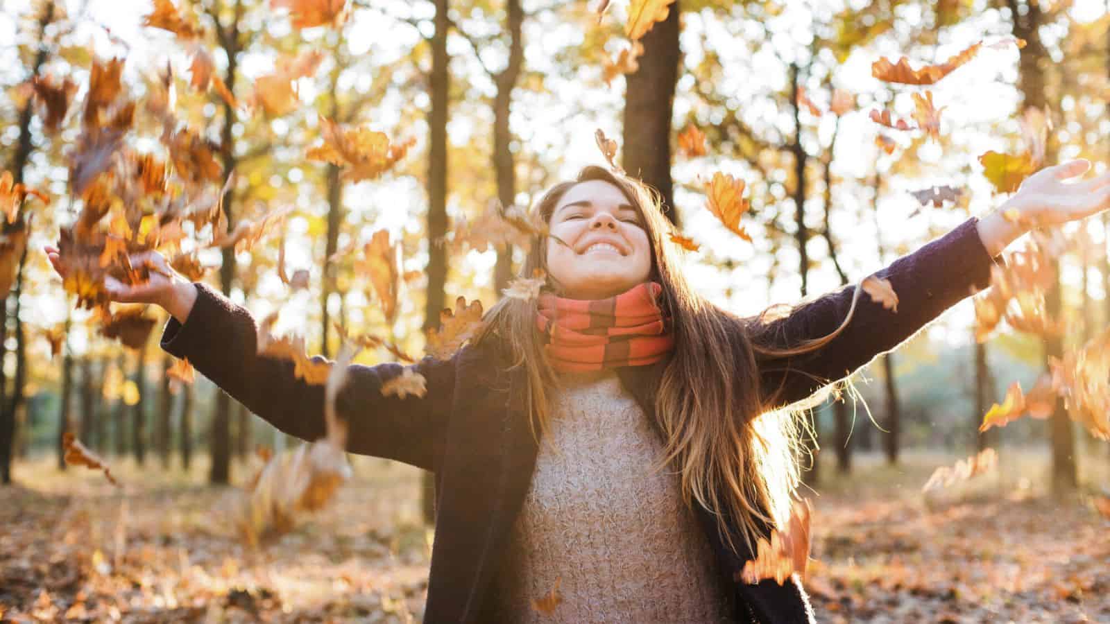 Woman smiling and throwing autumn leaves in the air in a sunlit forest.