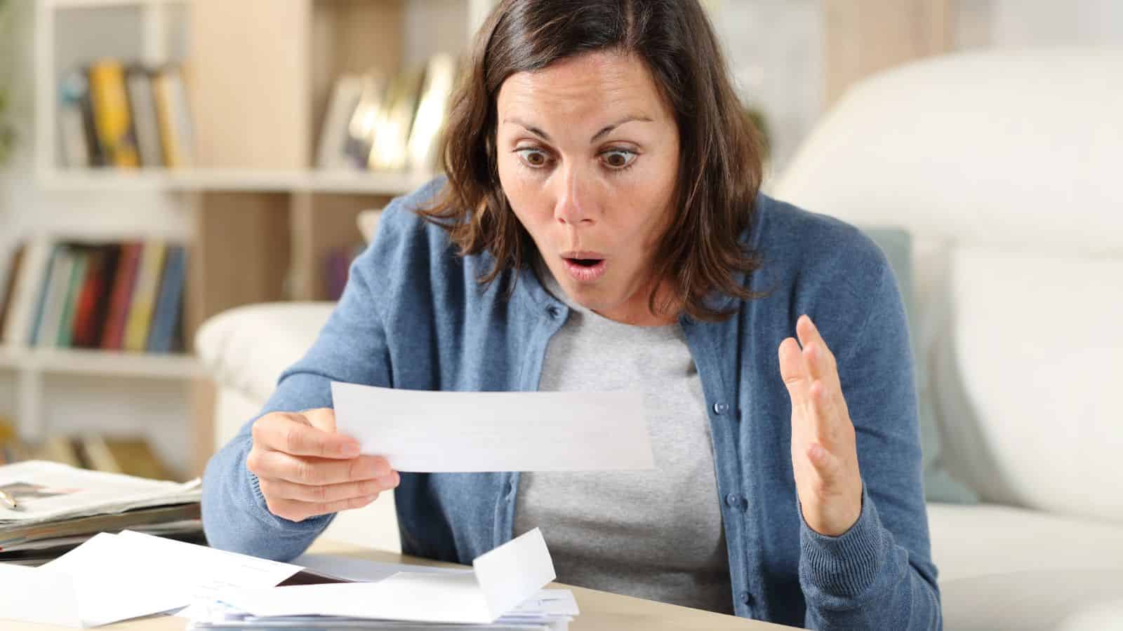 A woman looks surprised while reading a letter at a table with more papers and envelopes.