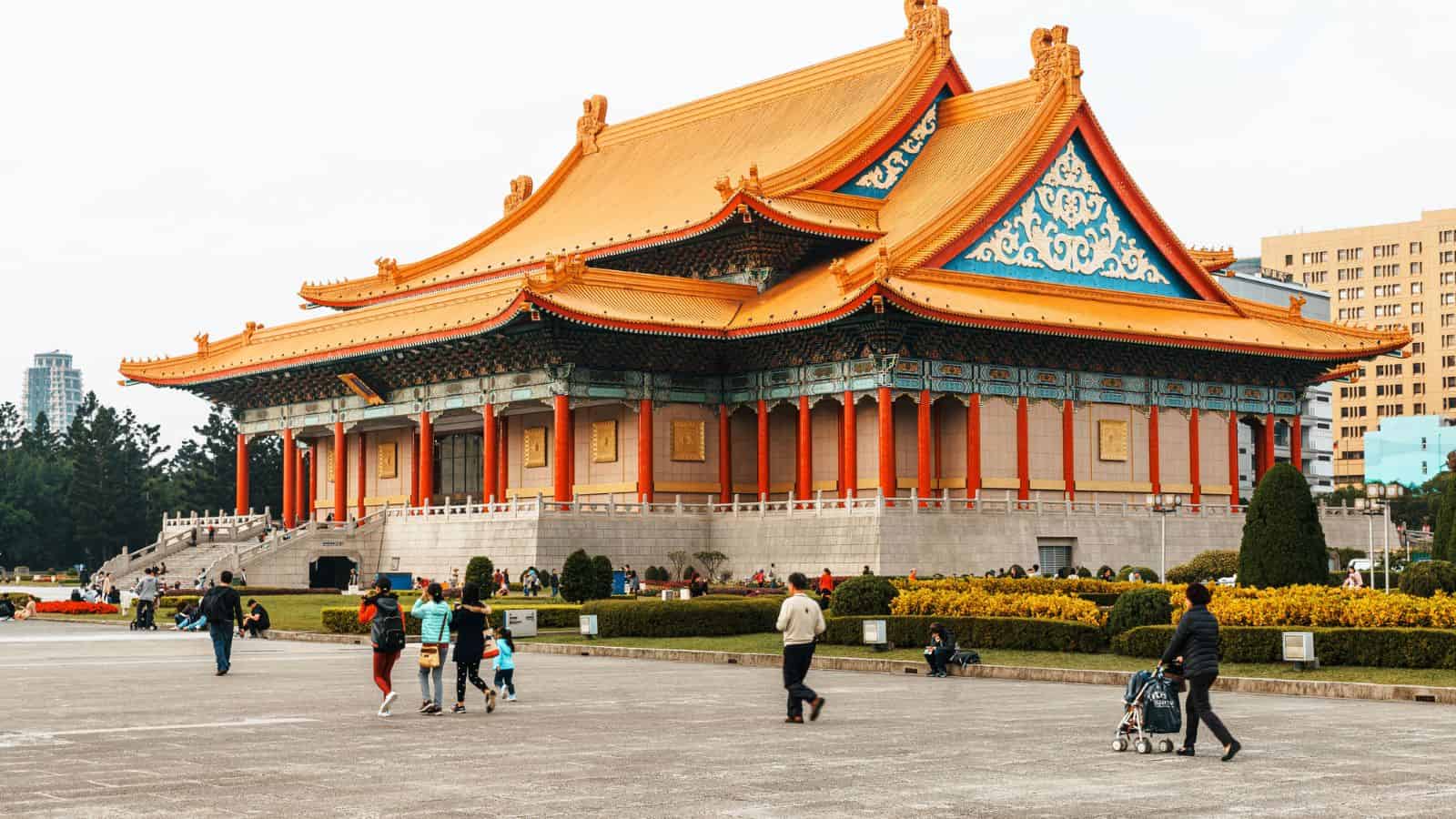 Large traditional Chinese building with an ornate orange roof, people walking in front in an open plaza.
