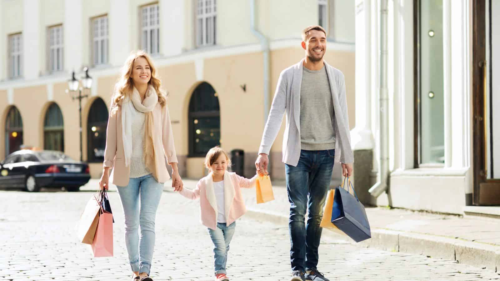 A smiling family of three walks on a city street holding shopping bags and hands on a sunny day.