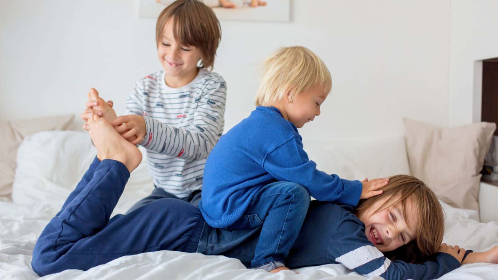 Three young children play and laugh together on a bed in a bright, cozy bedroom.