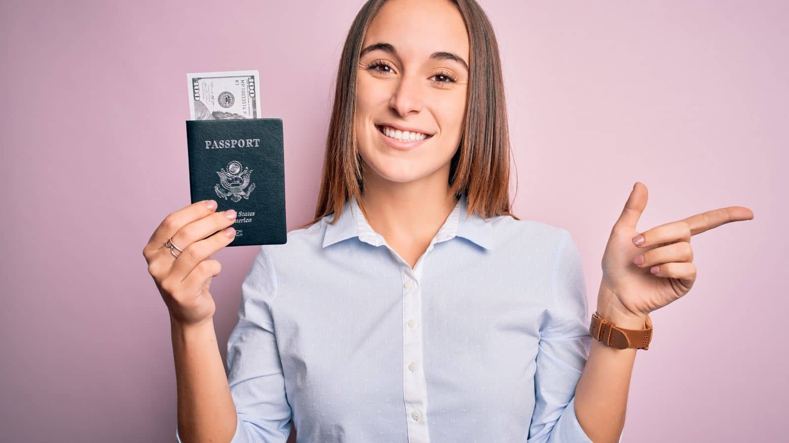 Smiling woman holding a U.S. passport with cash inside and pointing to the side against a pink background.