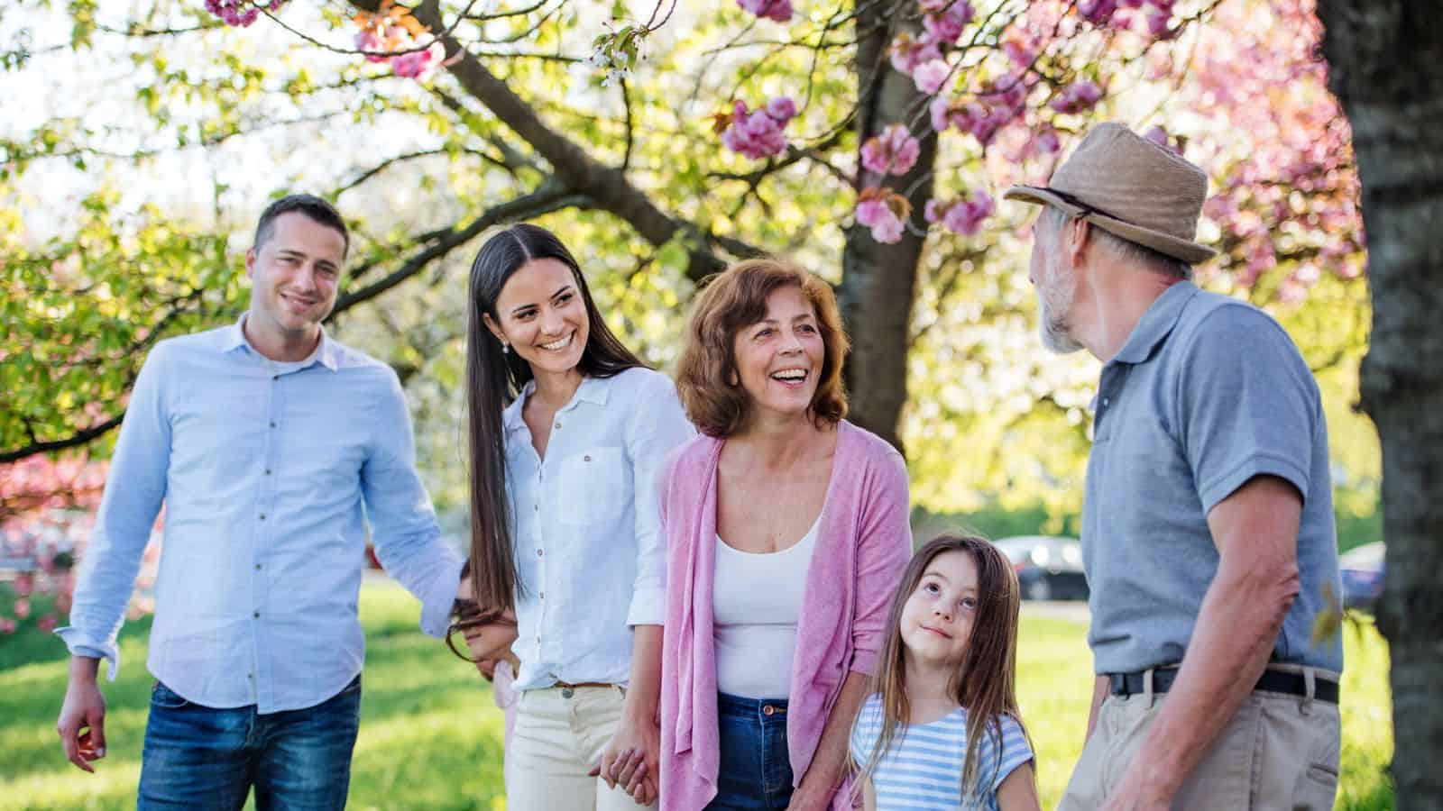 A family of five smiles and stands together outdoors under blooming trees on a sunny day.
