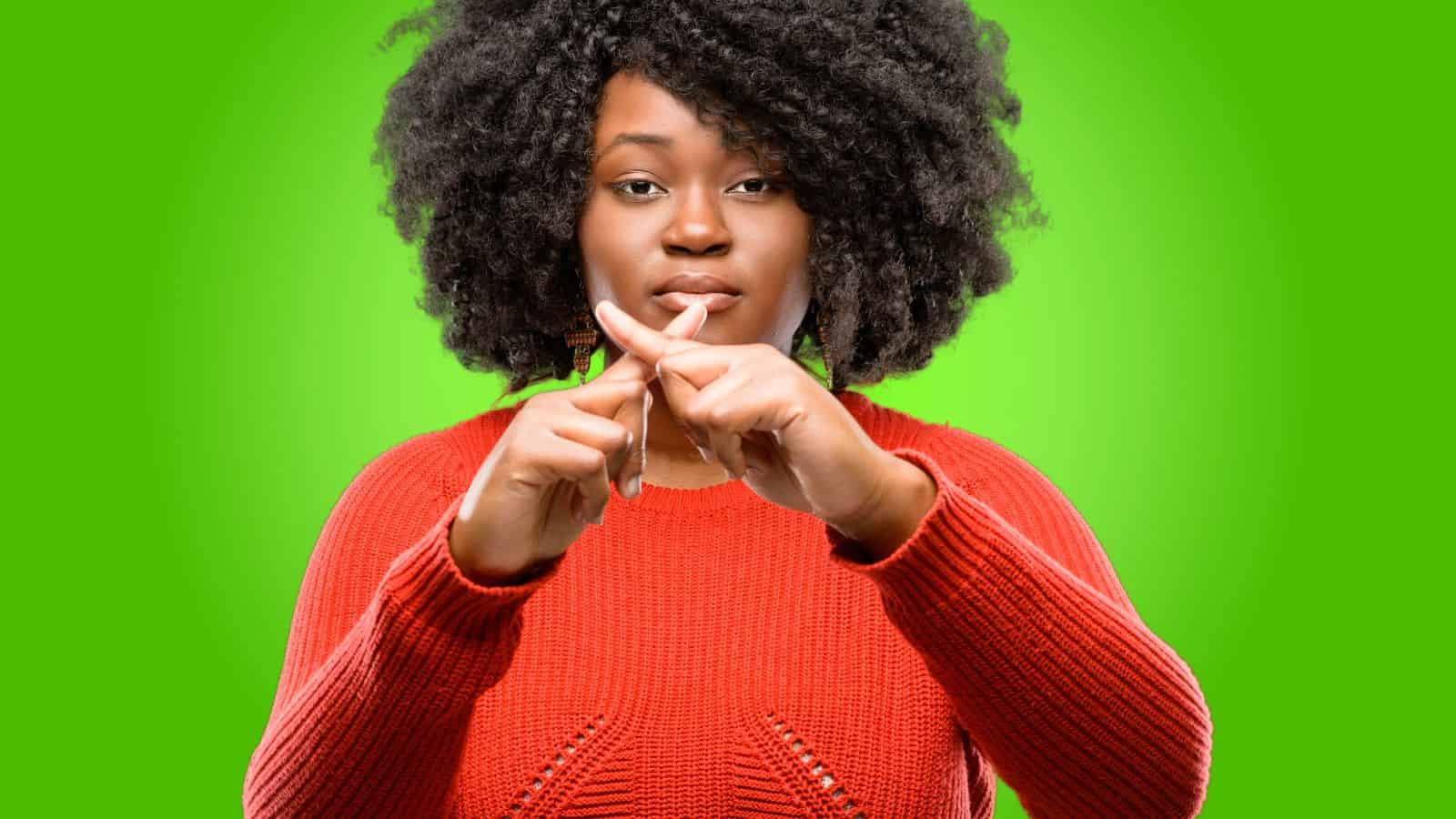 Woman in red sweater making an &ldquo;X&rdquo; with her fingers, signaling no, against a bright green background.